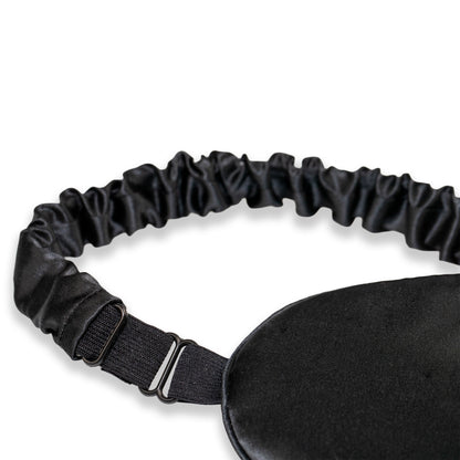 A close-up of a black SILKAVA silk sleep mask with an adjustable black elastic strap and metal slider buckle