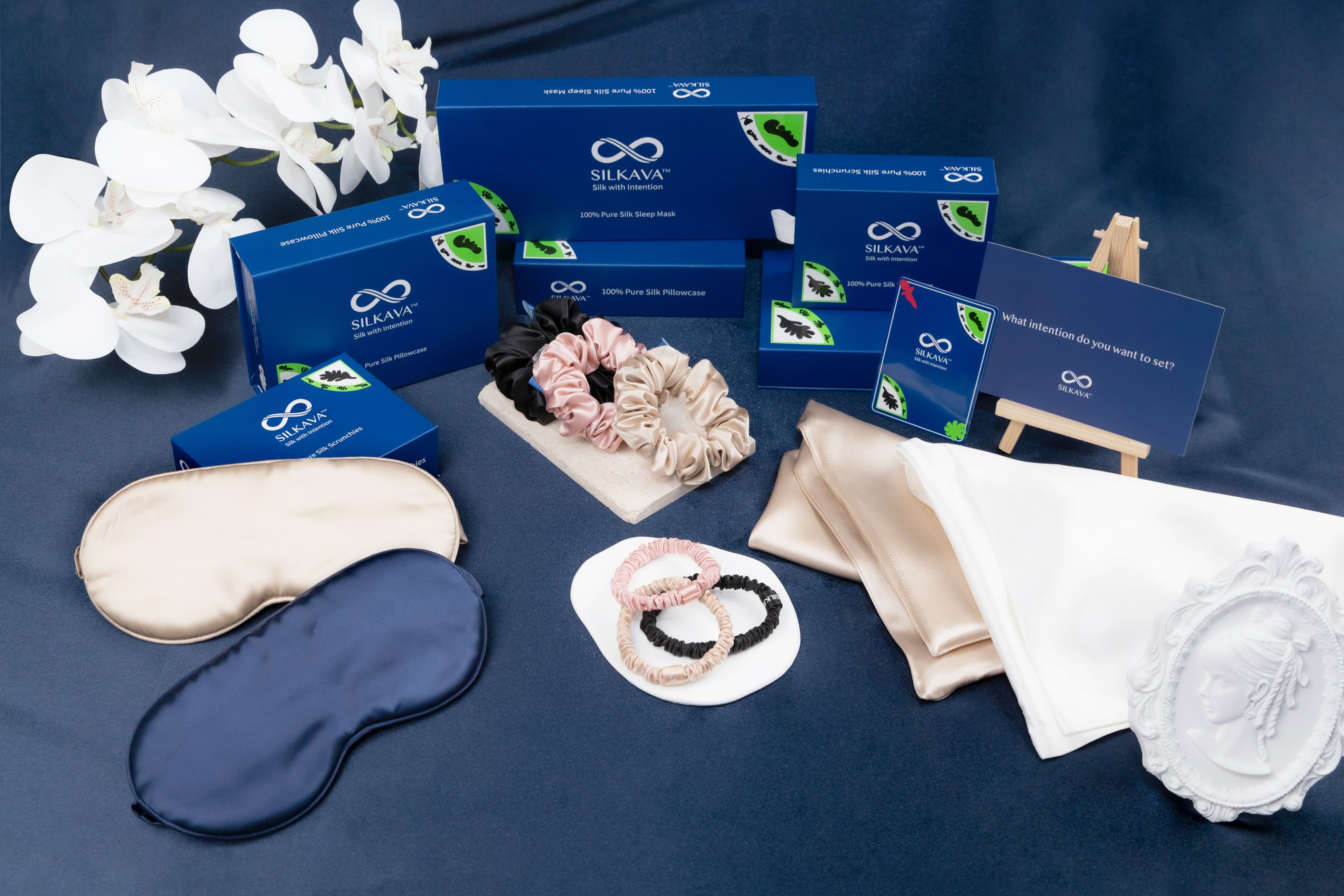 SILKAVA silk accessories and gift box packages, navy colorway lifestyle, including silk pillowcases, silk scrunchies, silk sleep masks in varied colors and sizes, and blue gift boxes with the SILKAVA logo and Origin Series design.